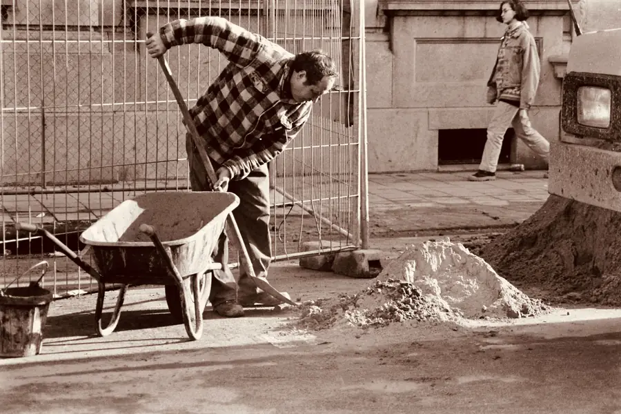 Paver shovelling sand into a wheelbarrow for a driveway foundation, sepia-toned black and white photo